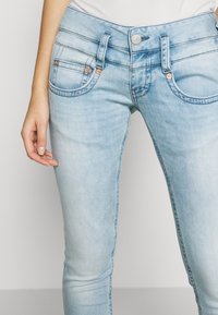 Hellblaue Denim-Jeans im Slim-Fit, mit kupferfarbenen Knöpfen, gesteppten Taschen und einer glatten Textur.