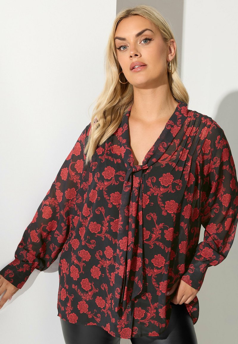 Sort bluse med rødt blomstermønster, lange ærmer, og en blød draperet halsudskæring. Stoffet virker let og flyder med en løs pasform.