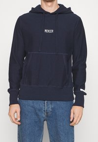 Sweat-shirt à capuche bleu marine en tissu doux avec une poche kangourou et un cordon de serrage. Présente "MERCER" brodé en blanc sur la poitrine.