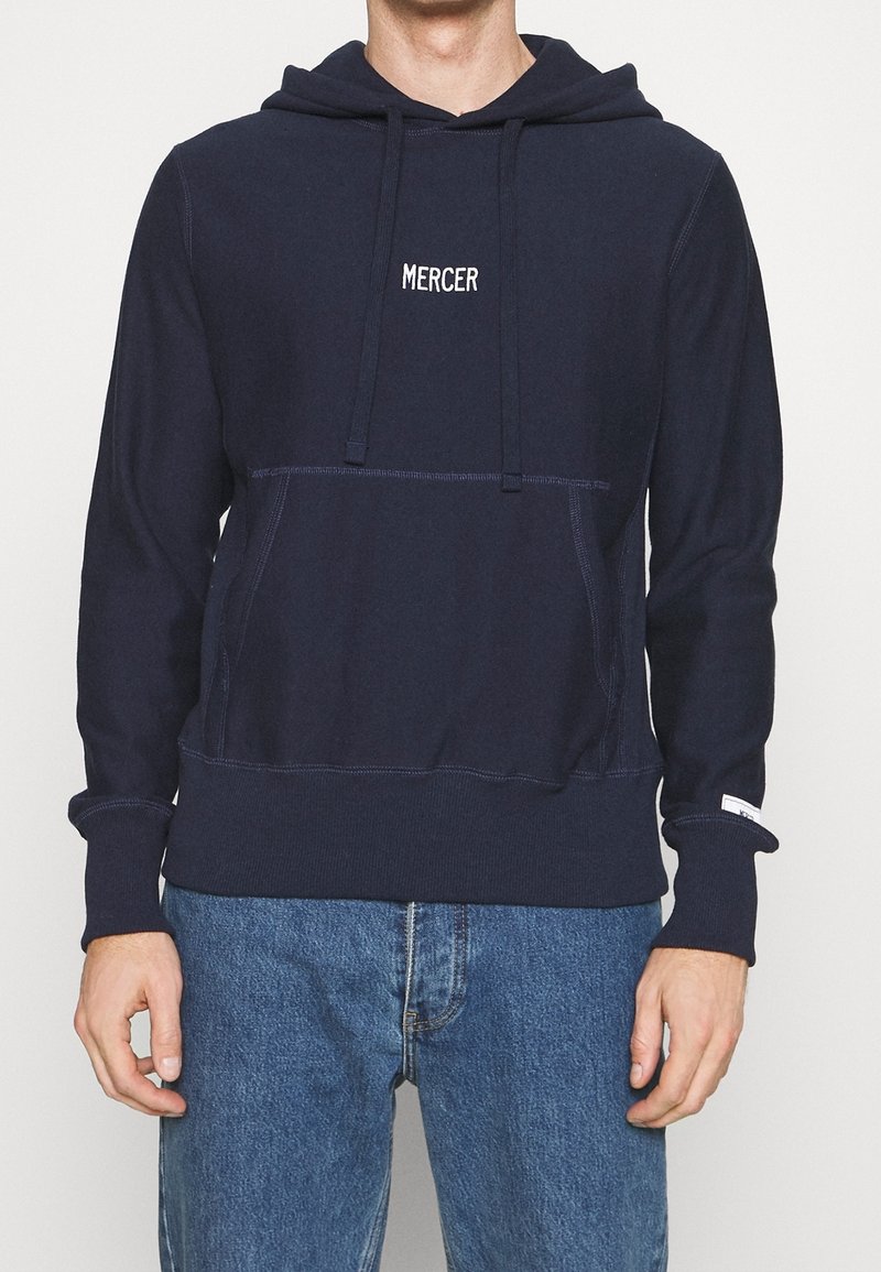 Sweat-shirt à capuche bleu marine en tissu doux avec une poche kangourou et un cordon de serrage. Présente "MERCER" brodé en blanc sur la poitrine.