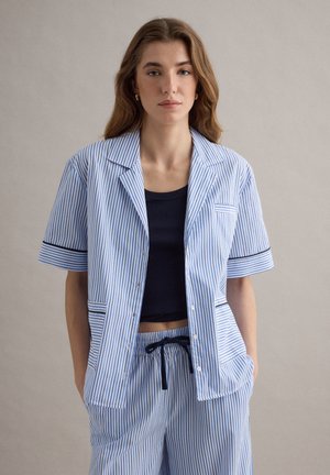 Junge Frau, die ein blau-weiß vertikal gestreiftes kurzärmeliges Hemd und passende Hosen mit schwarzem Kordelzug sowie ein schwarzes Tanktop trägt, steht.