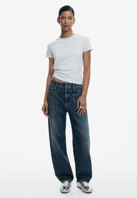 T-shirt ajusté à manches courtes de couleur bleu clair avec un logo subtil ; jean en denim bleu foncé large avec de grandes poches et une coupe décontractée ; baskets argentées.