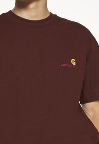 Burgundin puuvillapaita, jossa on pyöreä kaula-aukko ja lyhyet hihat. Paidassa on rintakehässä brodattu keltainen ja punainen Carhartt-logo.