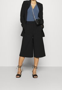 Culotte neri abbinati a un top blu sotto un blazer nero su misura. Gli accessori includono una piccola borsa a tracolla nera e sandali neri con cinturino.
