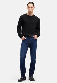 Czarny wełniany sweter, niebieskie jeansy denim, czarny skórzany pasek i czarne skórzane buty; model stoi z rękami w kieszeniach na jednolitym tle.