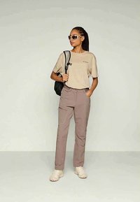 Kvinna med flätat hår som bär en beige t-shirt, mauve byxor, vita sneakers, solglasögon och en svart ryggsäck, stående mot en enfärgad bakgrund.