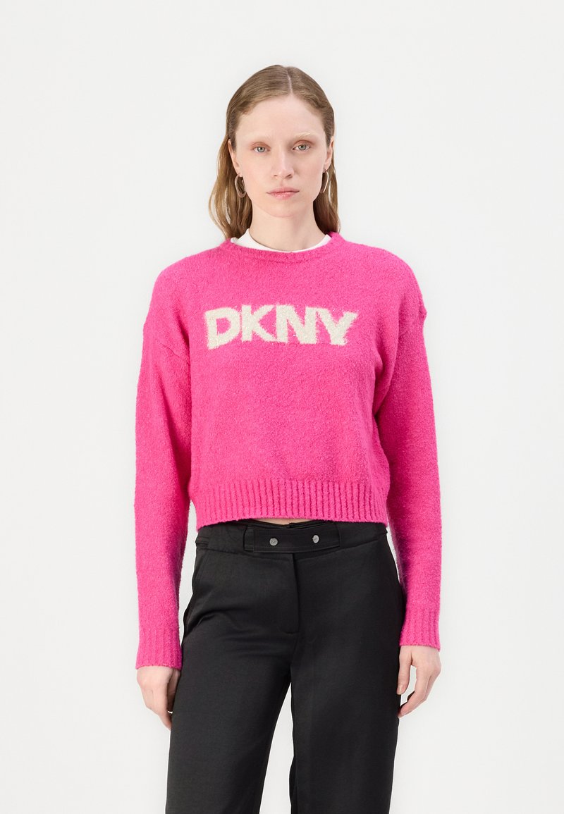 Vaaleanpunainen pörröinen lyhyt neule, jossa on valkoinen "DKNY"-logo, ribattu helma ja hihansuut, yhdistettynä mustiin housuihin. Tekninen materiaali tuo syvyyttä.