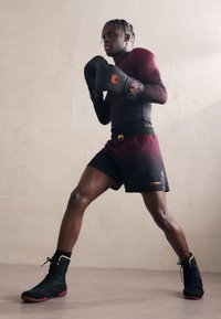 Αθλητής με μαύρη και μπορντό �εφαρμοστή μπλούζα και σορτσάκι, μαύρα γάντια πυγμαχίας και αθλητικά παπούτσια ψηλά, σε φως υφασμένο φόντο.