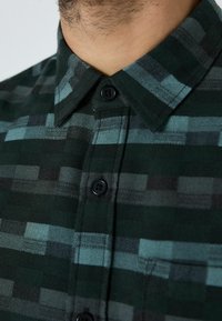 Vihreä ruudullinen paita, jossa on mustia ja teal-värisiä lohkoja, napitettava etuosa, peruskaulus sekä teksturoitu kangas näkyvillä ompelutiedoilla.