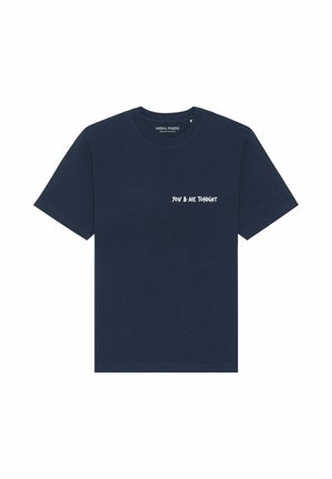 Jonge vrouw met lang bruin haar draagt een marineblauw oversized t-shirt met witte tekst "YOU & ME TONIGHT" op de borst.