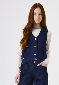 Dunkelblaues Jeans-Westen mit V-Ausschnitt, fünf Knöpfen und cropped Design. Kombiniert mit einem leichten, langärmligen Oberteil und dunklen Hosen.