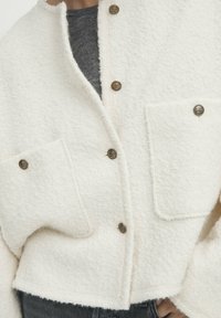 Biała, teksturowana, krótka kurtka z dużymi przednimi kieszeniami, złotymi guzikami i luźnym krojem, noszona na ciemnoszarej koszulce.