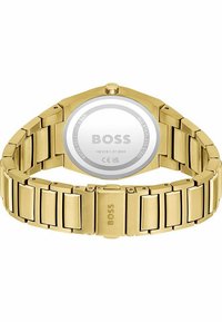 Reloj de acero inoxidable dorado con una caja octogonal, una pulsera texturizada y un fondo plateado que contiene los detalles de la marca y la resistencia al agua.