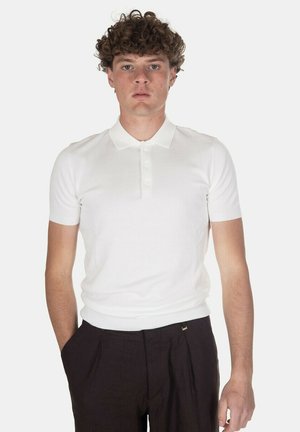Giovane uomo con capelli ricci che indossa una polo bianca aderente a maniche corte e pantaloni scuri plissettati, in piedi con le mani nelle tasche.