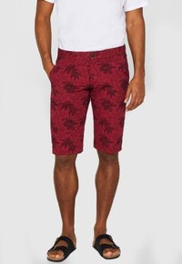 Homme portant un short rouge à motifs de palmiers noirs, un t-shirt blanc et des sandales noires à boucle, se tenant les mains dans les poches.