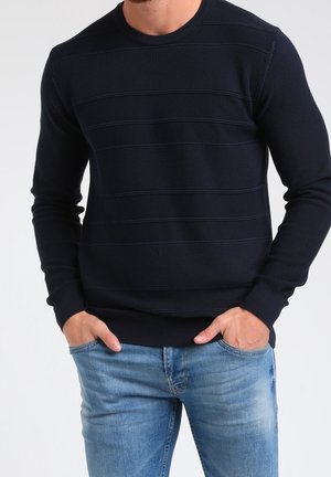 Mand iført navyblå tekstureret striktrøje med vandrette striber og lyseblå jeans, hænderne afslappet i lommerne.