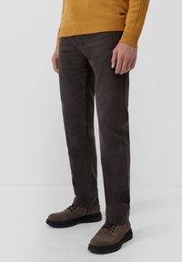 Muž v tmavohnedých džínsach, hnedých semišových šnurovacích topánkach a horčicovožltom pletenom svetri stojí na jednoliatom pozadí.