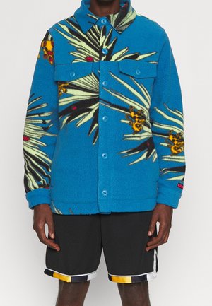 Niebieska kurtka polarowa z dużymi tropikalnymi liśćmi i kwiatowymi wzorami, z przodu zapinana na guziki i z klapkowymi kieszeniami na piersiach, noszona z czarnymi szortami z kolorowym obszyciem.