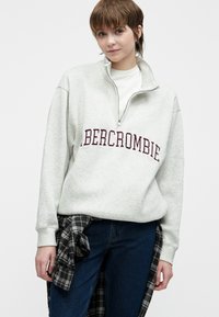 Młoda osoba w jasnoszarej bluzie rozpinanej na ćwierć z napisem "ABERCROMBIE" oraz ciemnoniebieskich dżinsach, w kratkowanej koszuli przewiązanej w pasie.