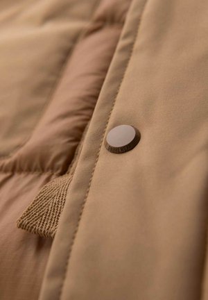 Beige Daunenjacke aus einem glatten Stoff, mit einem braunen Druckknopf, der "OMBREWEAR" beschriftet ist, und einem gewebten Etikett an der Seite.