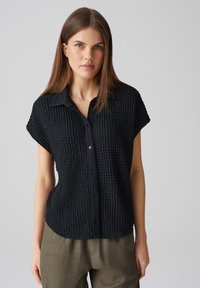 Camicia nera con texture, chiusura frontale con bottoni, maniche corte e colletto. Presenta un tessuto leggermente stropicciato e una vestibilità morbida.