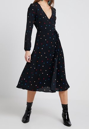 Kobieta w czarnej sukience kopertowej w wielokolorowe groszki, z długimi rękawami i błyszczącymi czarnymi botkami, stojąca na jednolitym tle.