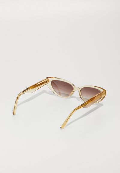 Gafas de sol estilo cat-eye con montura dorada transparente, lentes marrones degradados y patillas elegantes con un sutil acento textural.