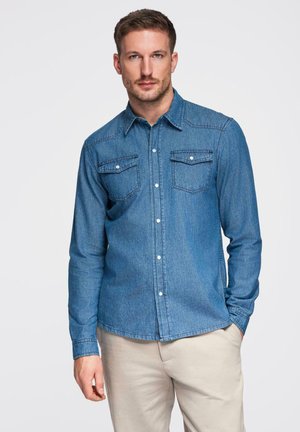 Jeansskjorta i mellanblått, med långa ärmar, knappar framtill och två bröstfickor med klaffar. Passar för vardagsbruk.