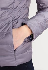 Jasnofioletowa, pikowana kurtka z gładkim wykończeniem, boczną kieszenią i widoczną podszewką w kropki przy krawędzi mankietu. Zamek błyskawiczny w kieszeni.