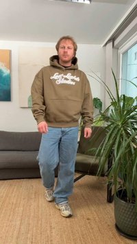 Pruun kapuutsiga dressipluus tekstiga "Love Light Studios", kombineeritud helesiniste teksade ja hallide tossudega, pehmes sisekeskkonnas taimedega.