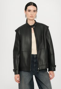 Giacca in pelle nera con colletto rialzato e chiusura con zip. Presenta maniche ampie, dettagli con cinturini regolabili e una texture liscia.