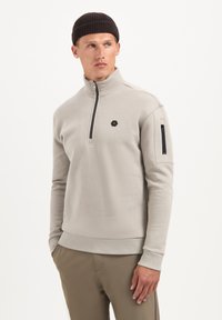 Lichtbeige pullover met een kwartritssluiting, gemaakt van een zachte stof met een geribde kraag. Voorzien van een logo op de borst en een ritszak op de linkermouw.