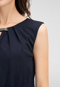 Marinblus utan ärmar med en plisserad design och en metallisk dekoration vid halsringningen, tillverkad av ett mjukt tyg med en slät struktur.