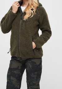 Chaqueta de forro polar verde oliva con un exterior texturizado, cierre de cremallera y dos bolsillos laterales. Cuenta con una capucha y cordones ajustables.