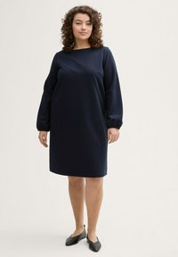 Marineblaue Kleid mit geradem Schnitt, langen, gepufften Ärmeln und rundem Ausschnitt. Der Stoff wirkt glatt mit einer leichten Textur.