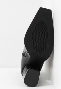 Chaussure de ville en cuir noir inclinée vers le haut, montrant une semelle texturée et un talon, placée sur un fond blanc d'angle.