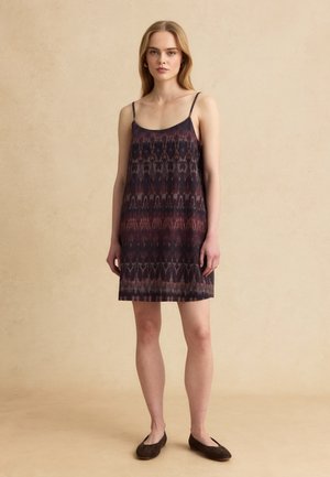 Giovane donna in piedi che indossa un vestito scuro senza maniche con fantasia, lungo fino al ginocchio, e ballerine scure su uno sfondo beige uniforme.