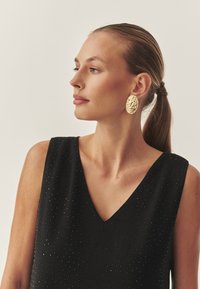 Ovalne uhančki z zlatim teksturiranim vzorcem, ki jih nosi model z repom. Črna brezaščitna majica ima globok V-izrez in pikčaste okraske.