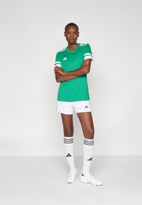 Groene sportt-shirt met witte strepen op de mouwen, gecombineerd met witte shorts en kniekousen met zwarte strepen. Sneakers zijn wit.