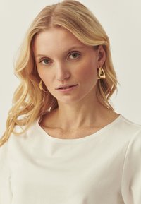 Top blanc à manches courtes avec un large décolleté, associé à des boucles d'oreilles triangulaires dorées. Cheveux blonds coiffés en ondulations lâches. Yeux verts visibles.