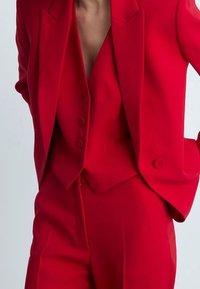 Costume rouge sur mesure avec une veste ajustée aux revers aiguisés et un gilet trois boutons en dessous, accompagné d'un pantalon slim.