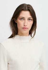 Suéter acanalado de color blanco roto con un cuello alto y ondulado. Textura suave, silueta ajustada y mangas largas. Diseño minimalista sin adornos.