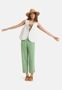 Chaleco acolchado blanco con un dobladillo redondeado, usado sobre una blusa marrón; combinado con pantalones de pierna ancha verde claro y sandalias de plataforma marrones.
