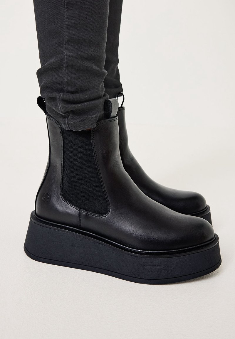 Bottes Chelsea à plateforme en cuir noir avec panneaux latéraux élastiques et languettes, portées avec un pantalon noir ajusté sur un fond neutre.