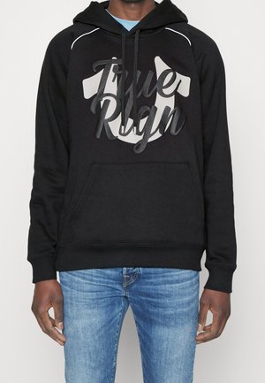 Man draagt een zwarte hoodie met de tekst "True Rign" en een schildlogo, gecombineerd met lichtblauwe spijkerbroek, staand tegen een effen achtergrond.