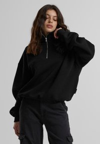 Svart zip-up hettegenser med vide ermer og ribbede mansjetter, med en subtil logo på brystet og en løs passform. Båret med mørke cargobukser.