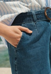High-waisted blue denim jeans featuring a fitted design, side pockets, and a textured waistband with a large round belt buckle.