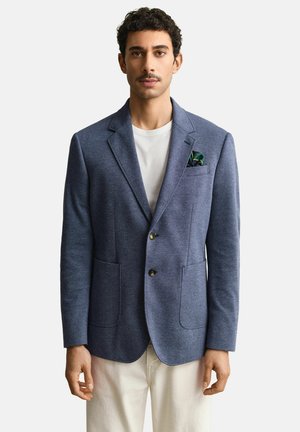Man draagt een blauw colbert met een groen pochet, een wit overhemd en beige broek, staand tegen een effen achtergrond.