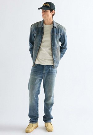 Młody mężczyzna w dżinsowej kurtce, wyblakłych niebieskich dżinsach, beżowych butach i czarnej czapce, stojący z rękami za plecami, patrzący na bok.