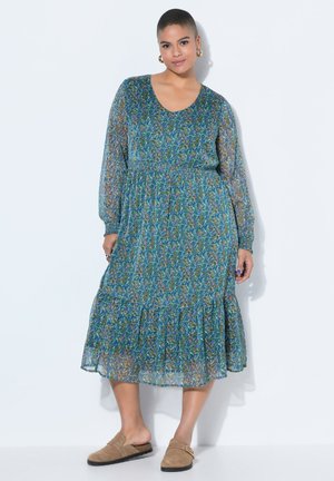 Frau steht und trägt ein blaues, langärmeliges, knielanges Kleid mit Blumenmuster und braune Slipper, vor einem schlichten weißen Hintergrund.